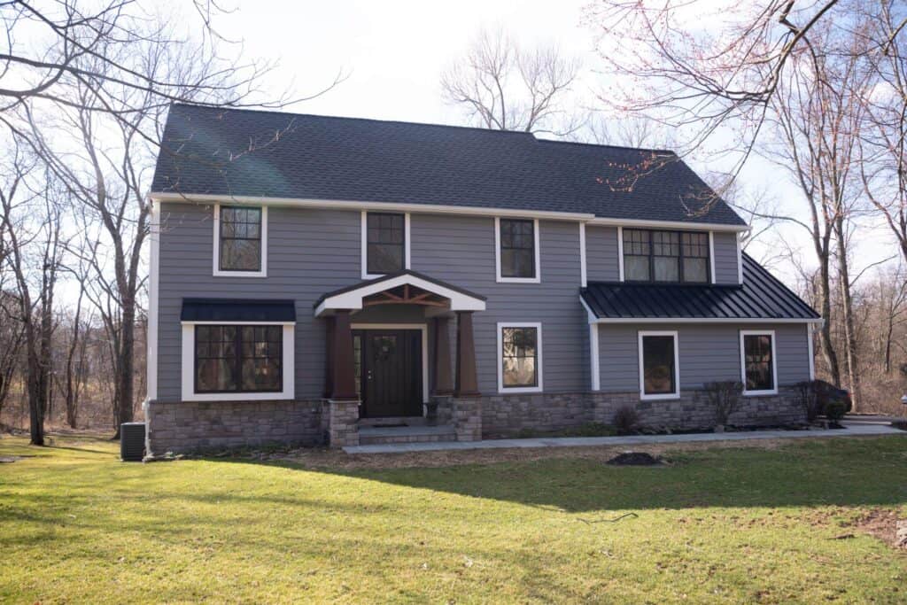 a two story gray house with white trim and black roof, featuring a stone lower facade and a covered front entrance, surrounded by leafless trees and a grassy yard on a sunny day.