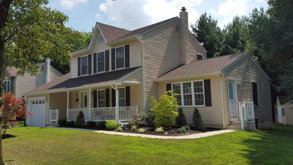 a beige two story suburban house with white trim, a covered front porch, dark shutters, attached garage, and a well maintained lawn with landscaped bushes and trees.
