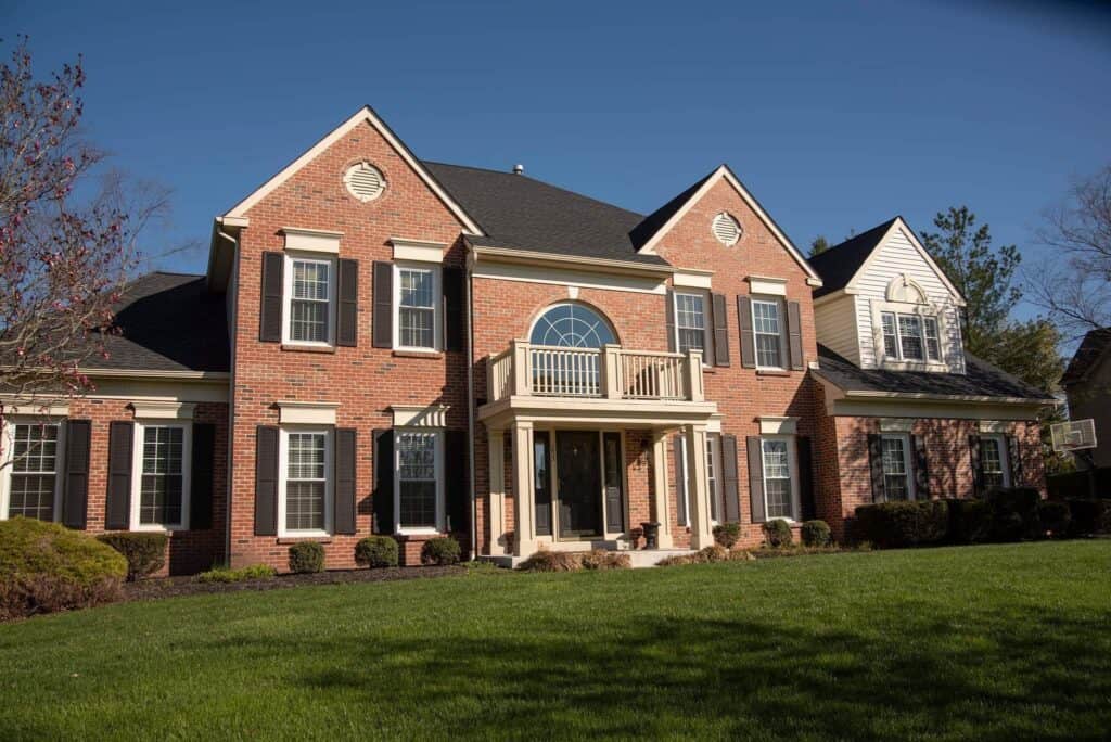 two story brick house with black shutters, white trim, and a balcony above the front entrance, set on a well kept lawn under a clear blue sky.