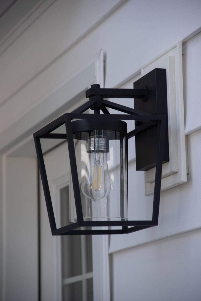 a modern black metal outdoor wall lantern with a clear glass cylinder and visible light bulb is mounted on the white siding of a house near a door.