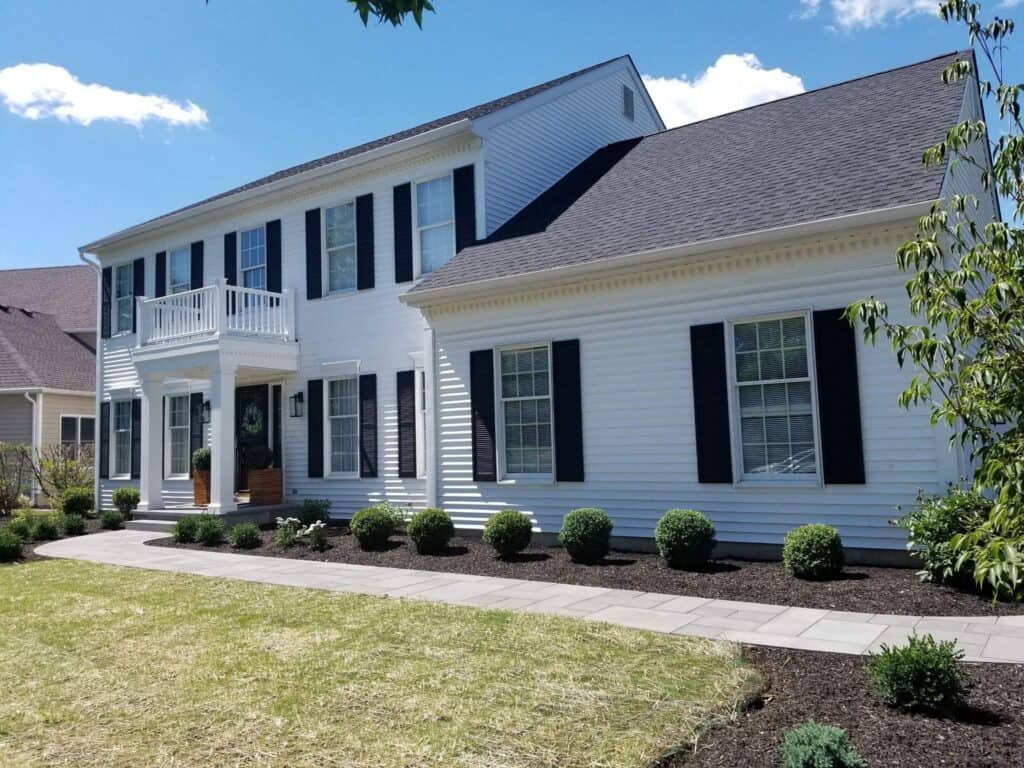 a white two story house with black shutters, a front porch with columns, and neatly trimmed bushes along a paved walkway under a sunny, blue sky.