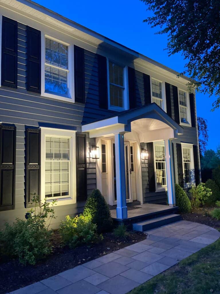 a two story house with gray siding, white trim, and black shutters is lit up at dusk. the front door is framed by columns and a small porch, surrounded by neatly trimmed bushes and a stone walkway.