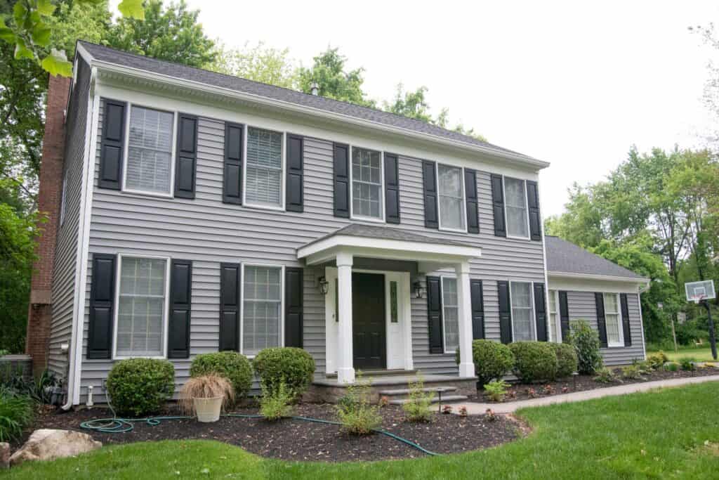 a two story gray house with black shutters and a covered front porch, surrounded by neatly trimmed bushes and a landscaped yard. a basketball hoop is visible in the background on the right.