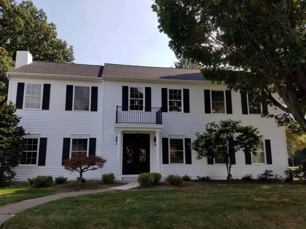 a two story white house with black shutters and a black front door, featuring a small balcony above the entrance, surrounded by trees, bushes, and a neatly trimmed lawn.