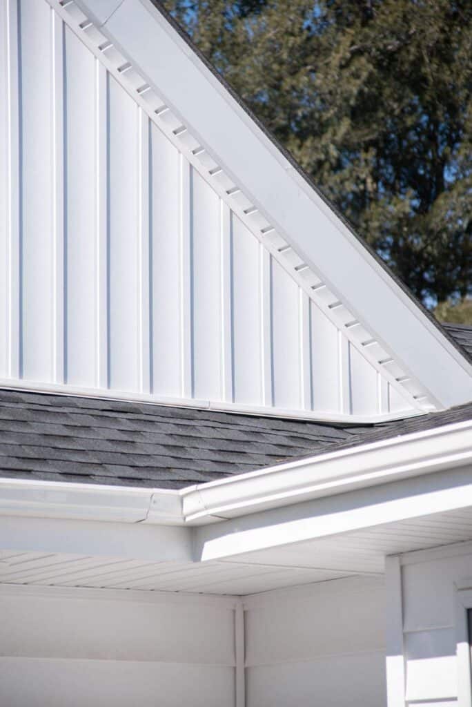 close up of a house roof with black shingles, white vertical siding, and white trim, showing the intersection of rooflines and part of a gutter, with trees blurred in the background.
