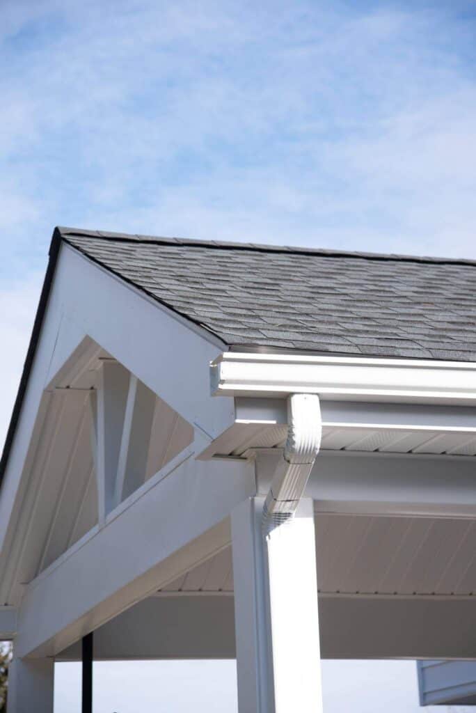 a close up view of the corner of a white outdoor structure with a dark shingled roof, white gutter, and downspout, against a partly cloudy blue sky.