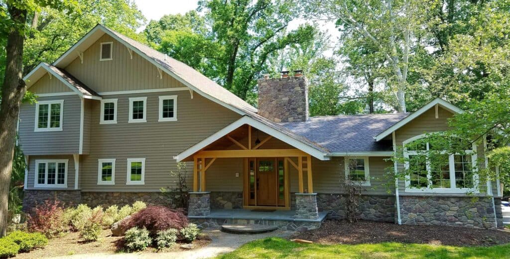 a two story house with beige siding, white trim, and a stone chimney. the front porch features wooden beams, and the house is surrounded by trees, shrubs, and green lawn.