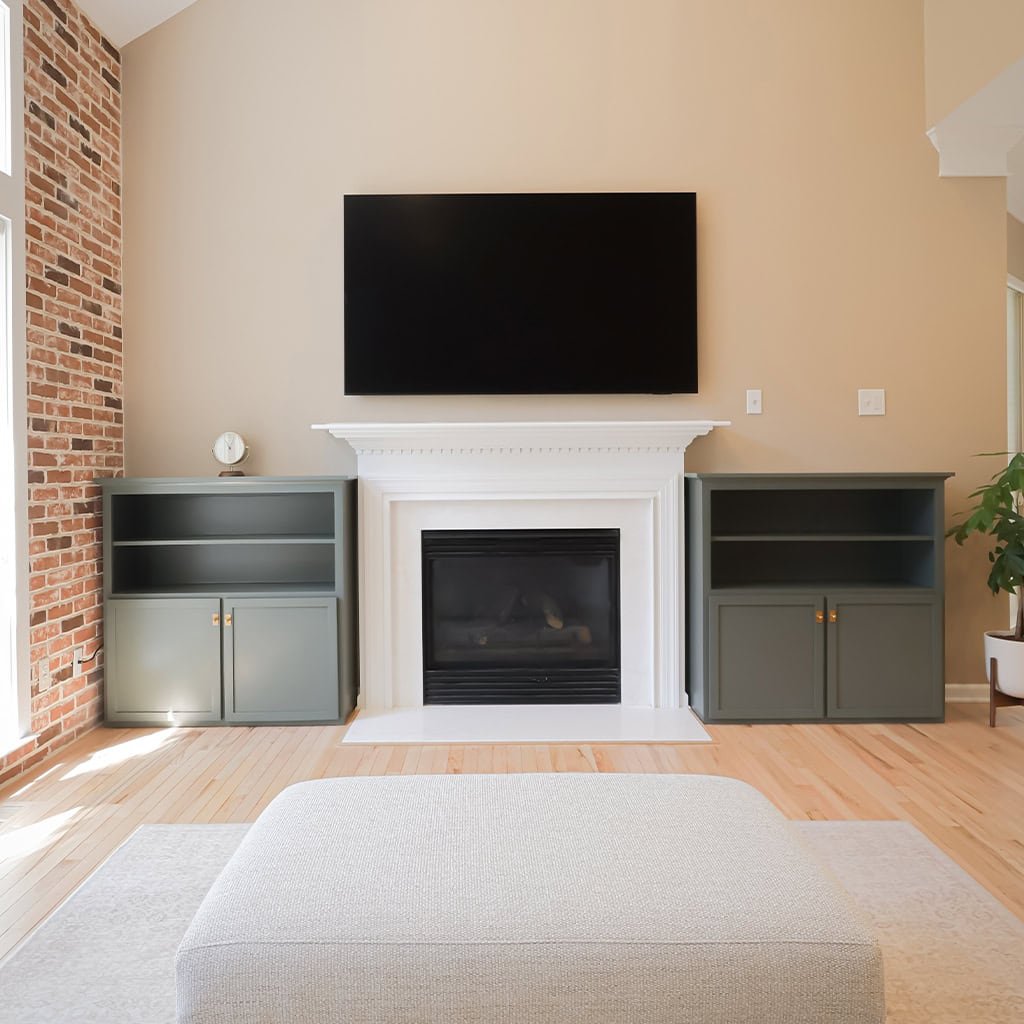 a living room with a brick accent wall, a black fireplace with a white mantel, a large flat screen tv above it, two green built in cabinets on either side, and a light ottoman in the foreground. a living room with a brick accent wall, a black fireplace with a white mantel, a large flat screen tv above it, two green built in cabinets on either side, and a light ottoman in the foreground.