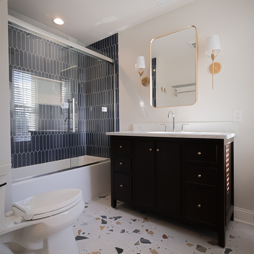 modern bathroom with a dark vanity, gold fixtures, a white toilet, and a bathtub with a glass door. the shower features dark blue tile, and geometric patterned floor tiles add visual interest. modern bathroom with a dark vanity, gold fixtures, a white toilet, and a bathtub with a glass door. the shower features dark blue tile, and geometric patterned floor tiles add visual interest.