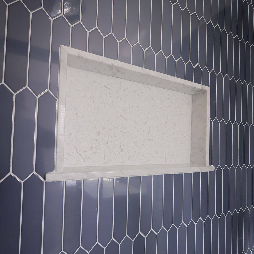 a recessed white marble shower niche with a floral pattern inside is set into a wall of glossy blue elongated hexagon tiles arranged vertically. a recessed white marble shower niche with a floral pattern inside is set into a wall of glossy blue elongated hexagon tiles arranged vertically.