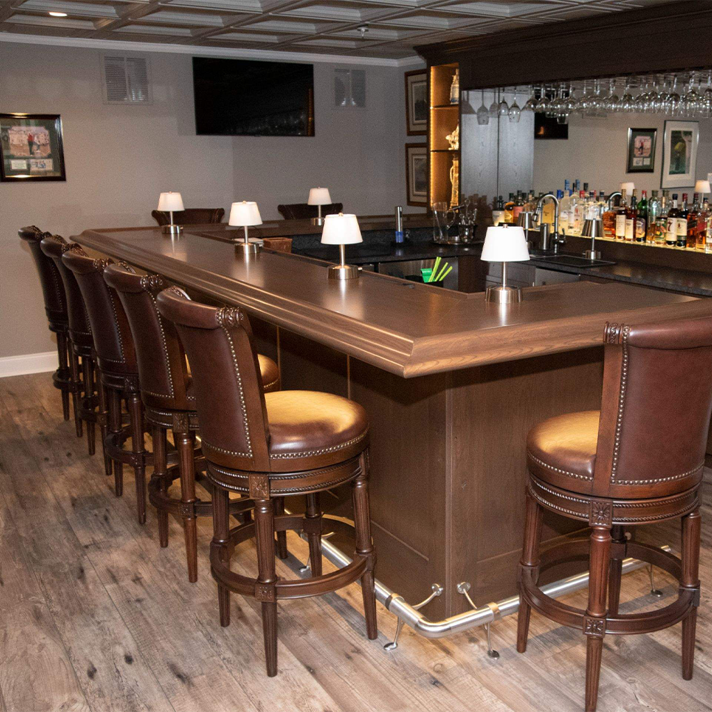 a home bar with brown leather barstools, a wooden counter, bottles and glasses on shelves, small lamps on the bar, and wood flooring—a perfect addition to any kitchen remodeling bucks county project. framed pictures hang on the beige walls in the background.