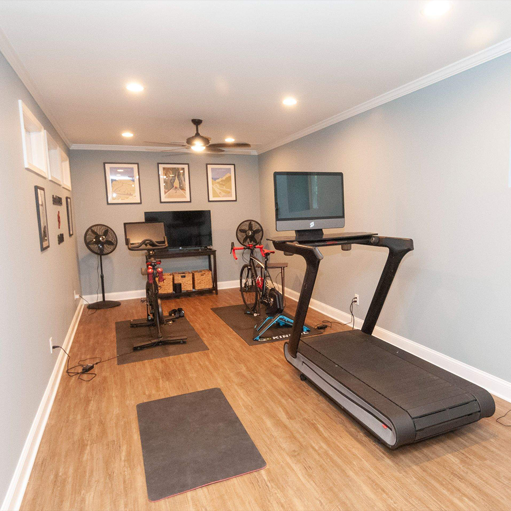home gym with a treadmill, two bike trainers, two floor fans, a tv, and framed pictures on gray walls—perfect inspiration while planning your next project. the room has wood flooring and ceiling lights, much like the modern style seen in recent kitchen remodeling bucks county transformations.