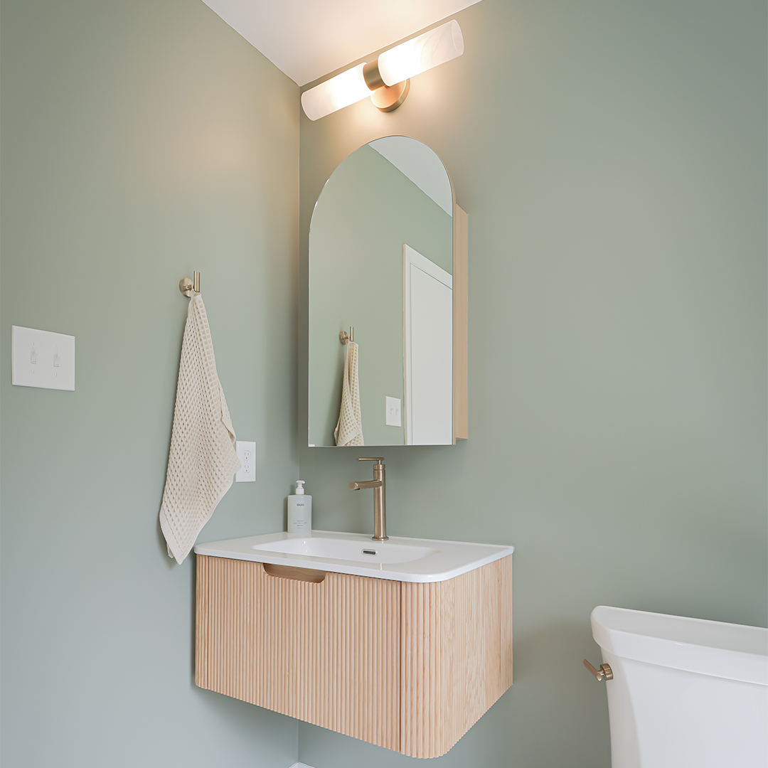 modern bathroom with sage green walls, a floating wood vanity with a white sink, an arched mirror, gold faucet, wall towel hook with beige towel, soap dispenser, and a white toilet partially visible. modern bathroom with sage green walls, a floating wood vanity with a white sink, an arched mirror, gold faucet, wall towel hook with beige towel, soap dispenser, and a white toilet partially visible.