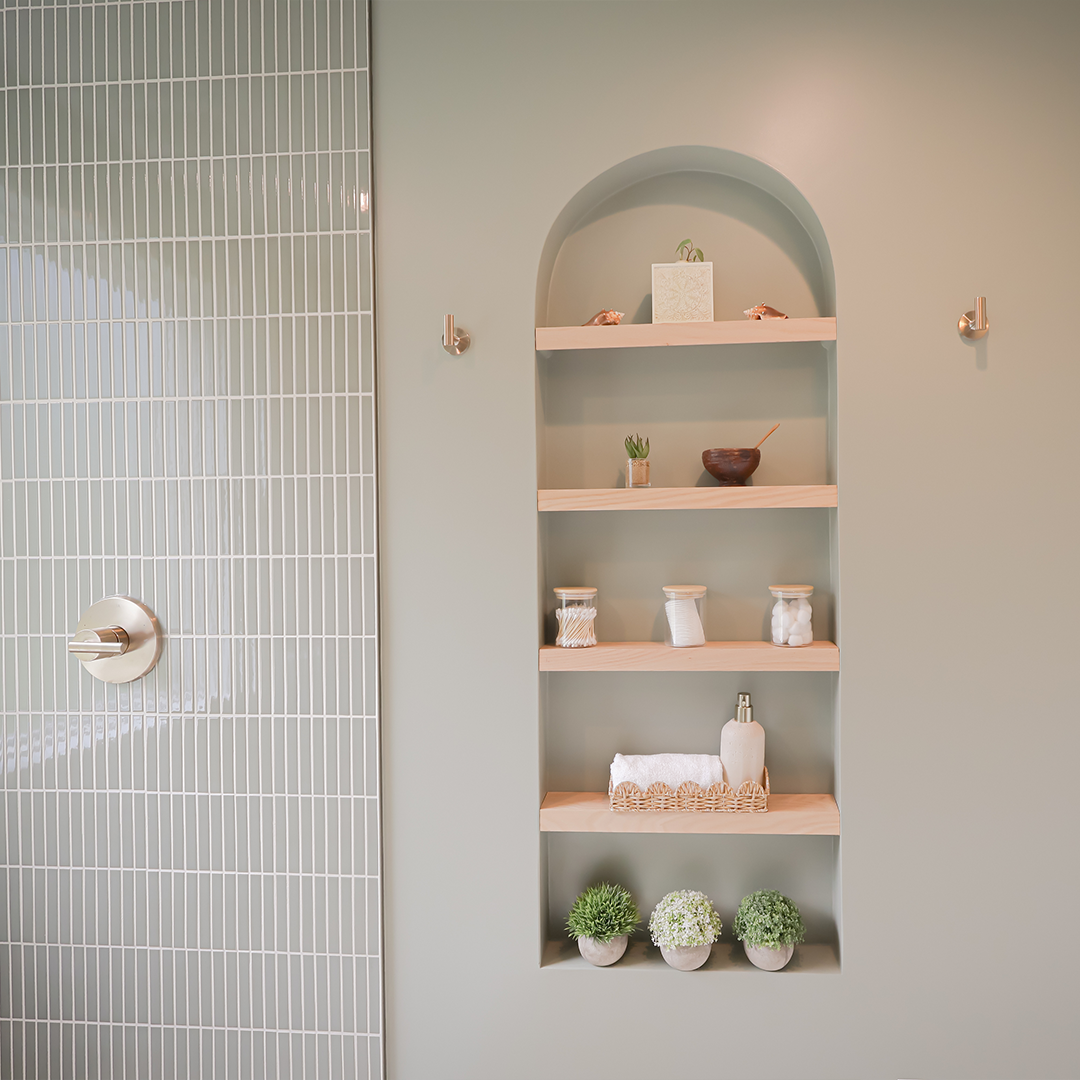 built in arched shelf with decorative items, towels, jars, and plants on wood shelves; nearby is a tiled shower wall with a silver faucet. the wall and shelf are painted a soft, muted green. built in arched shelf with decorative items, towels, jars, and plants on wood shelves; nearby is a tiled shower wall with a silver faucet. the wall and shelf are painted a soft, muted green.