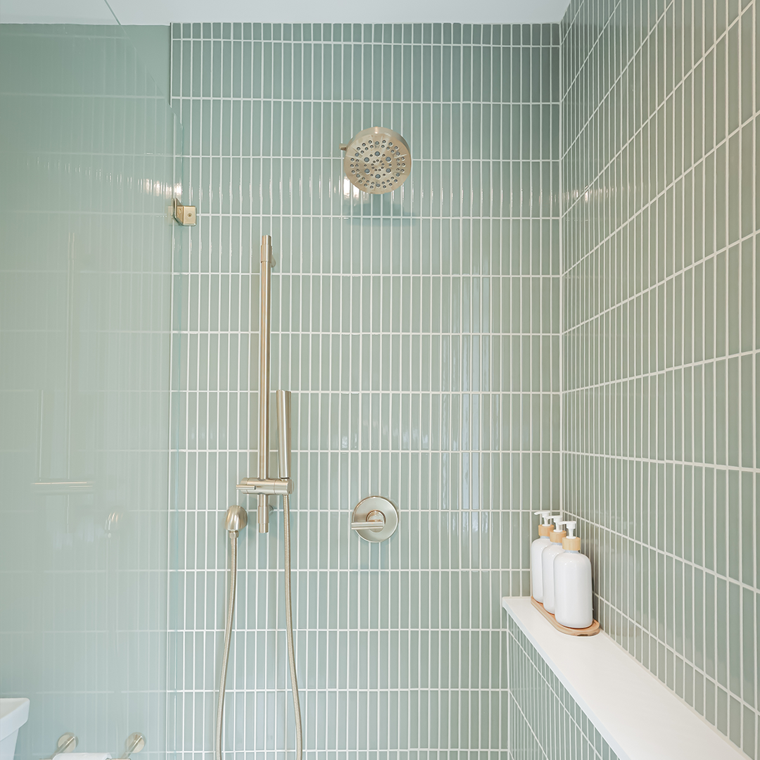 modern shower with light green vertical subway tiles, a silver rain shower head, matching fixtures, and three bottles on a white shelf. the glass shower door is partially visible on the left side. modern shower with light green vertical subway tiles, a silver rain shower head, matching fixtures, and three bottles on a white shelf. the glass shower door is partially visible on the left side.