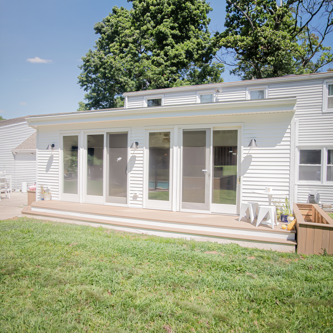 a modern white house with large glass doors, small porch steps, a planter box, white stools, and green lawn; trees and blue sky are in the background. a modern white house with large glass doors, small porch steps, a planter box, white stools, and green lawn; trees and blue sky are in the background.