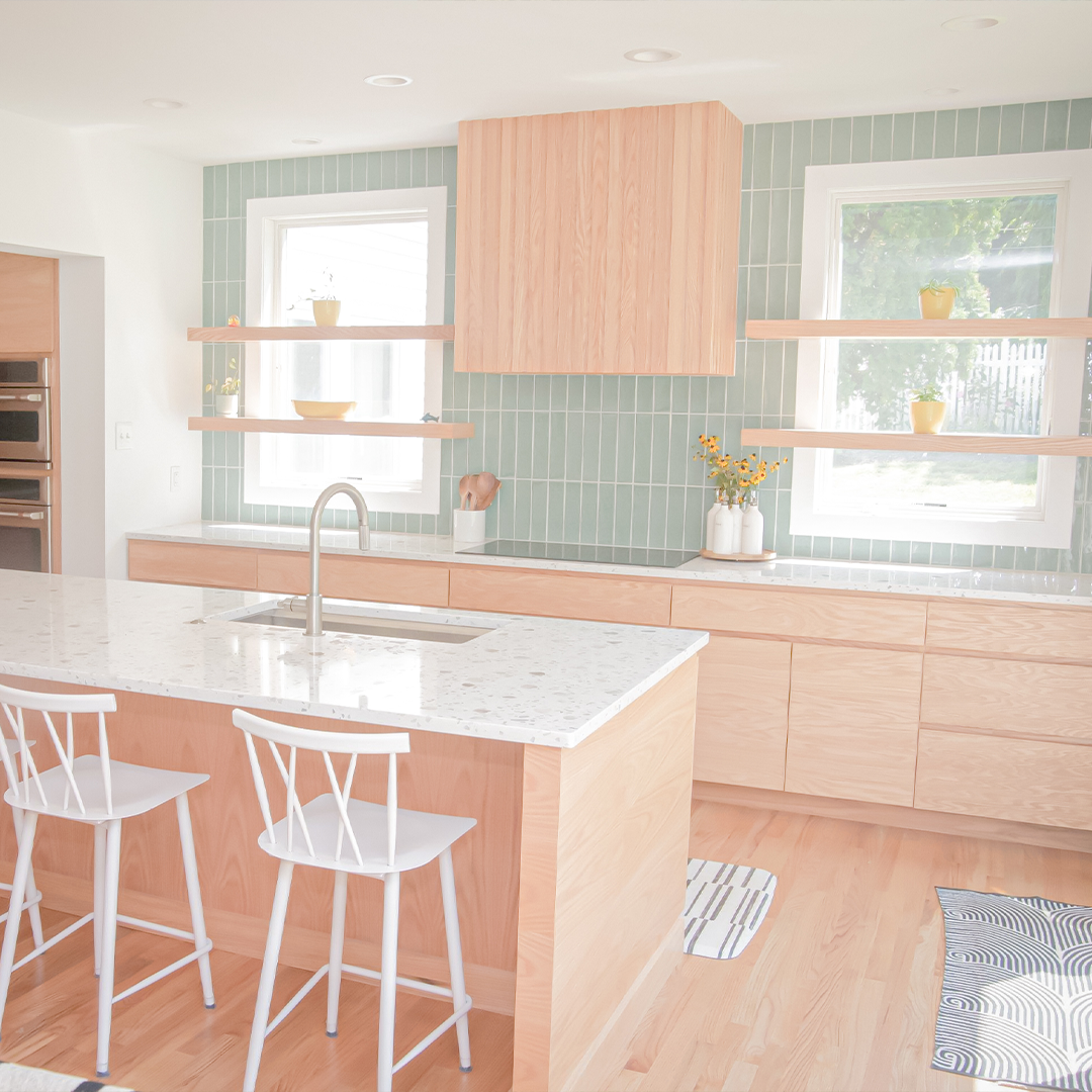 bright, modern kitchen with light wood cabinets, white countertops, an island with two white chairs, green tile backsplash, open shelves with yellow décor, and large windows letting in natural light. bright, modern kitchen with light wood cabinets, white countertops, an island with two white chairs, green tile backsplash, open shelves with yellow décor, and large windows letting in natural light.