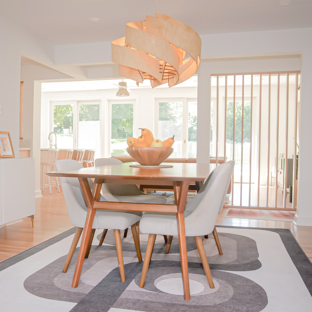 modern dining area with a wooden table, four light upholstered chairs, a fruit bowl centerpiece, a unique spiral pendant light, and large windows letting in natural light. a geometric rug covers the light wood floor. modern dining area with a wooden table, four light upholstered chairs, a fruit bowl centerpiece, a unique spiral pendant light, and large windows letting in natural light. a geometric rug covers the light wood floor.