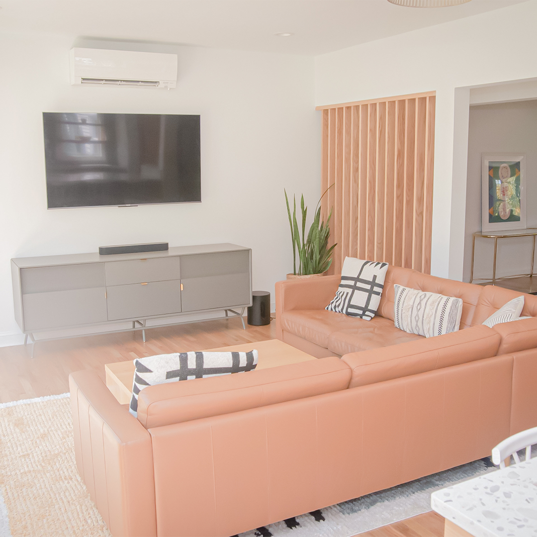modern living room with a tan leather sectional sofa, patterned pillows, a large wall mounted tv, a gray console, green plant, and wooden accent slats. light wood floors and minimalistic decor create a bright, airy feel. modern living room with a tan leather sectional sofa, patterned pillows, a large wall mounted tv, a gray console, green plant, and wooden accent slats. light wood floors and minimalistic decor create a bright, airy feel.