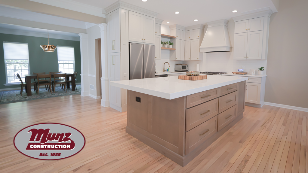 modern kitchen with white cabinets, stainless steel appliances, a large island with drawers, and light wood flooring. the adjacent dining area offers inspiration for those exploring sunroom ideas or planning four season sunrooms in bucks county. munz construction logo in lower left corner. bright, open space.