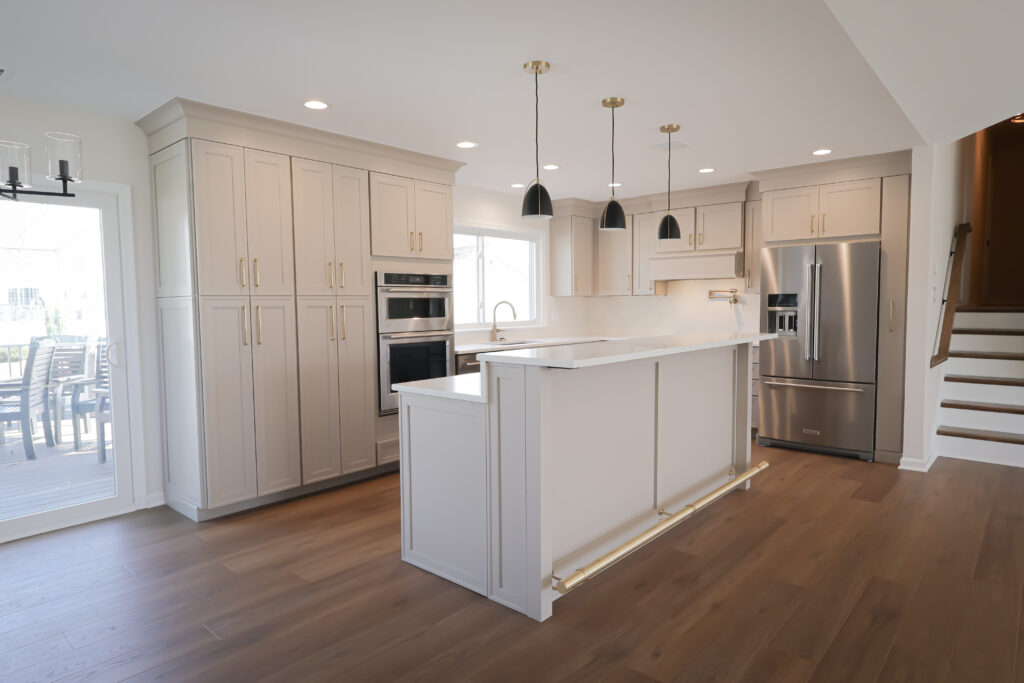 modern kitchen with white cabinets, stainless steel appliances, a center island with gold accents, pendant lights above, wood flooring, and a view of a patio through glass doors on the left—perfect inspiration if you’re exploring sunroom ideas or considering adding a four season sunroom to your home.