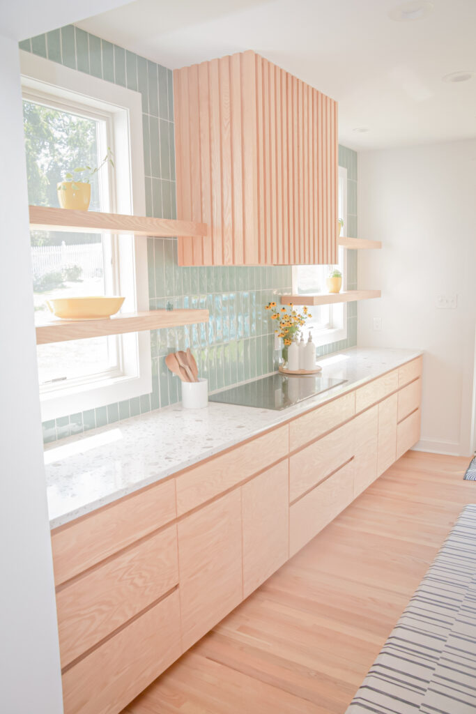 bright, modern kitchen with light wood cabinets, open shelves, green tile backsplash, white countertop, and natural light from two windows—a stunning example of kitchen renovation bucks county style. a vase with flowers and dishes decorate the shelves and countertop.