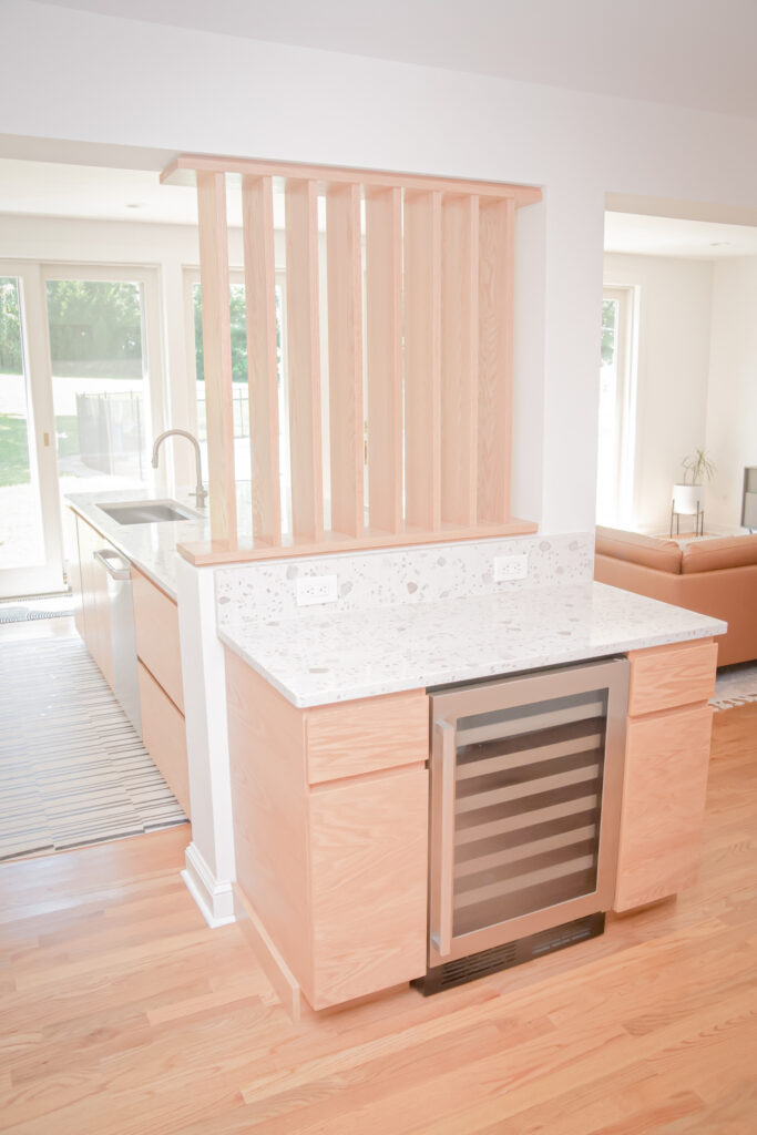 a modern kitchen with light wood cabinets and floors, a white countertop, a built in wine fridge, a partition with vertical slats, and large windows letting in natural light—this space perfectly showcases the possibilities of kitchen renovation bucks county for creating stylish and functional interiors.