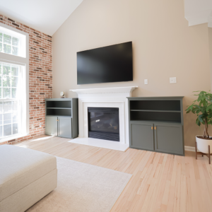 a living room with a fireplace, two green cabinets on each side, a wall mounted tv above the fireplace, a brick accent wall, hardwood floors, and a potted plant in the corner.