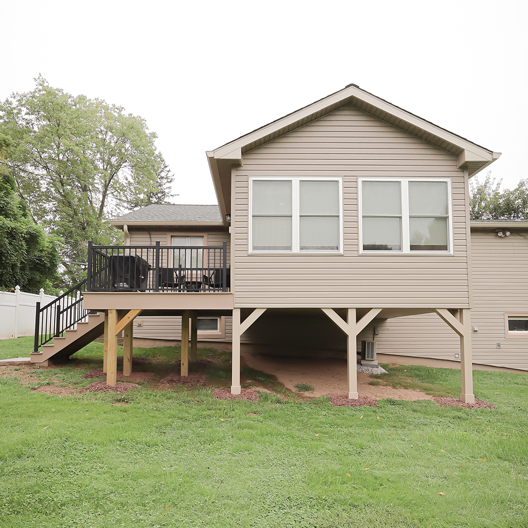 a beige house with a raised sunroom addition on stilts, multiple windows, and an adjacent deck with black railings, set on a grassy lawn with some trees in the background. a beige house with a raised sunroom addition on stilts, multiple windows, and an adjacent deck with black railings, set on a grassy lawn with some trees in the background.