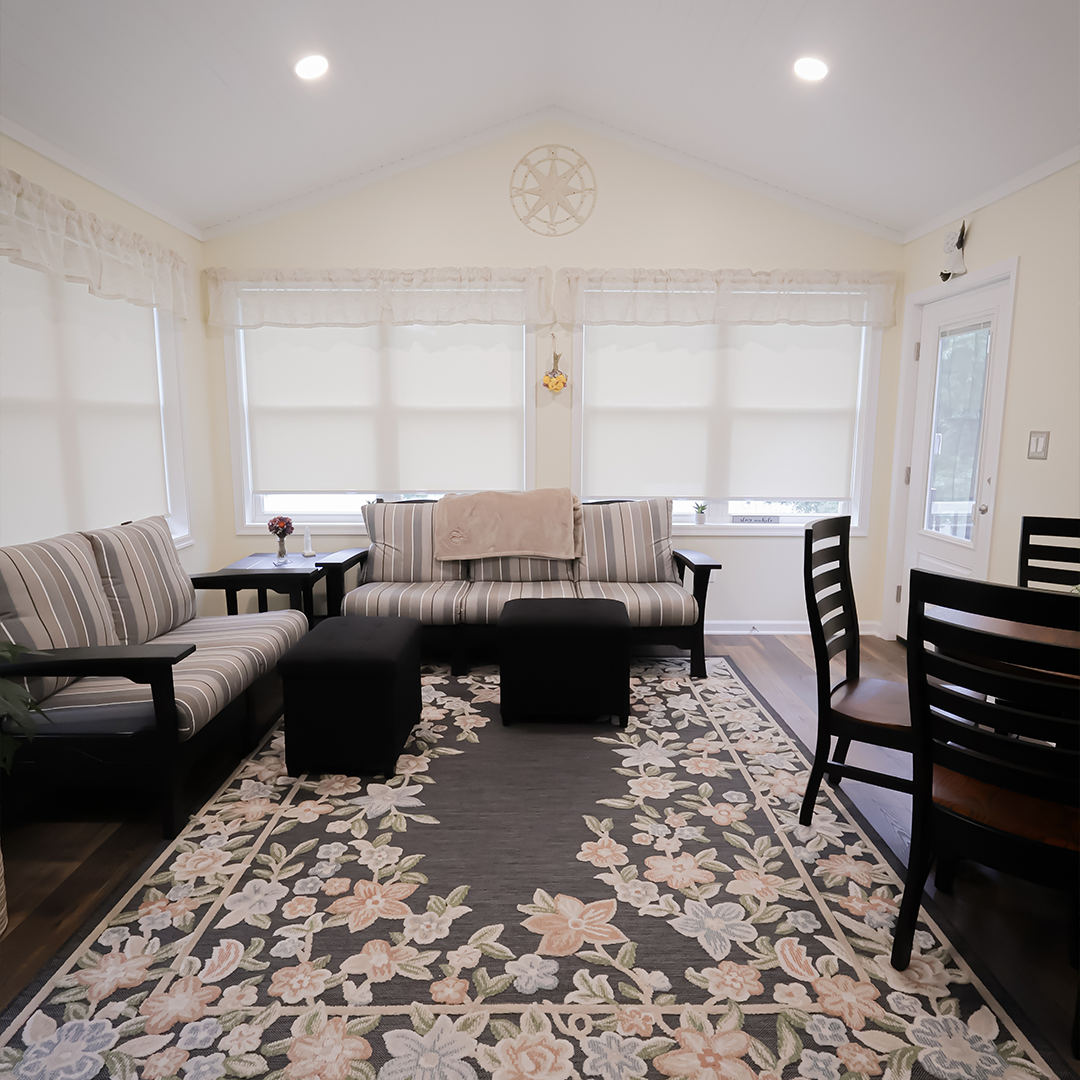 bright living room with two striped sofas, a floral rug, two black ottomans, a round dining table with chairs, and large windows with white blinds. the walls and ceiling are light colored. bright living room with two striped sofas, a floral rug, two black ottomans, a round dining table with chairs, and large windows with white blinds. the walls and ceiling are light colored.