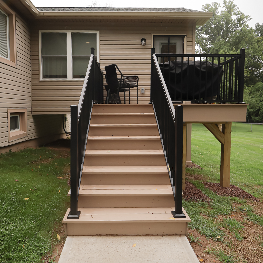 stairs lead up to a small deck attached to a beige house, with black railings, a grill, and patio furniture. the deck overlooks a grassy yard with trees in the background. stairs lead up to a small deck attached to a beige house, with black railings, a grill, and patio furniture. the deck overlooks a grassy yard with trees in the background.