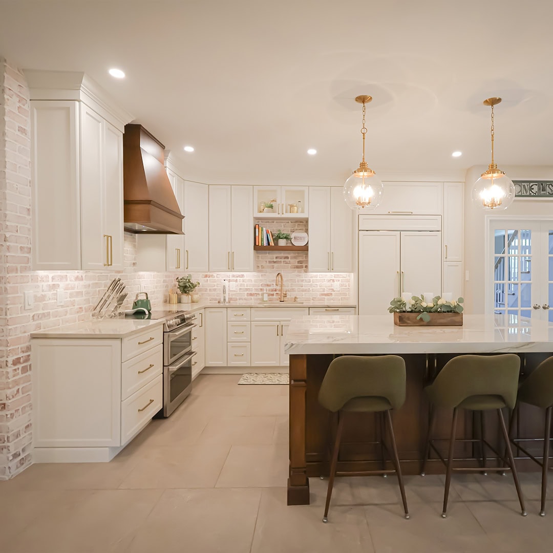 White kitchen with a brick accent wall, wooden range hood, large center island with green stools, and two gold pendant lights. White kitchen with a brick accent wall, wooden range hood, large center island with green stools, and two gold pendant lights.