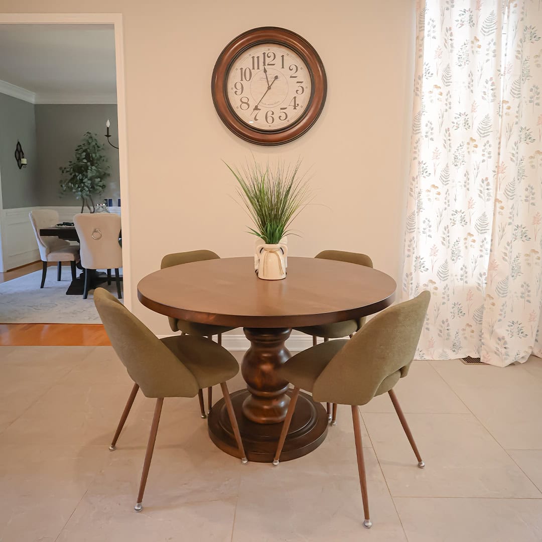 A round wooden pedestal table with four olive green chairs, topped with a potted plant, centered under a large wall clock. A round wooden pedestal table with four olive green chairs, topped with a potted plant, centered under a large wall clock.
