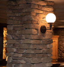 a round wall sconce with a glowing white bulb is mounted on a rustic stone pillar inside a dimly lit room with wooden furniture in the background, creating an inviting atmosphere perfect for your next kitchen renovation bucks county project.